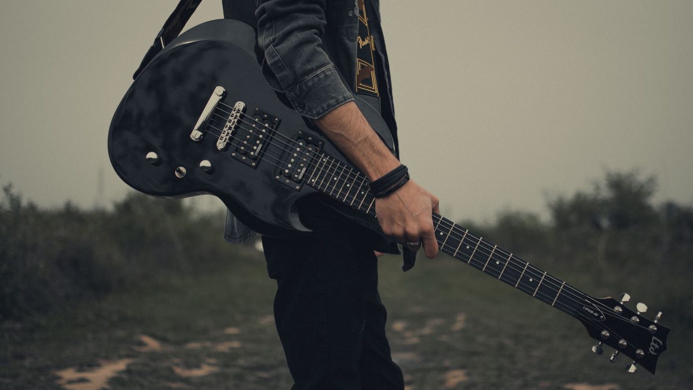 A young man holding a black electric guitar standing in an open field. Musician, outdoor rock style.