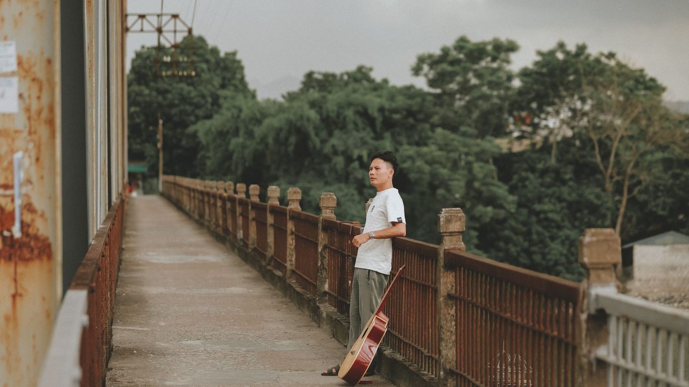 A man stands with a guitar on a bridge, surrounded by lush greenery.
