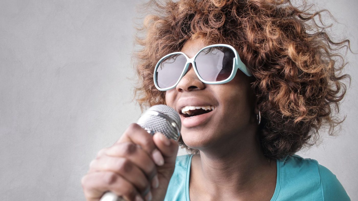 Close-up of a young woman with curly hair and sunglasses singing passionately into a microphone.