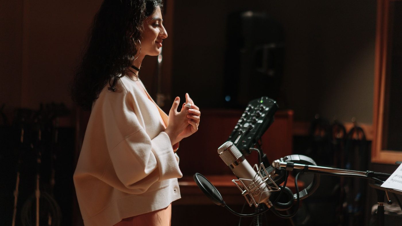 Side view of a woman singing into a condenser microphone in a professional recording studio.