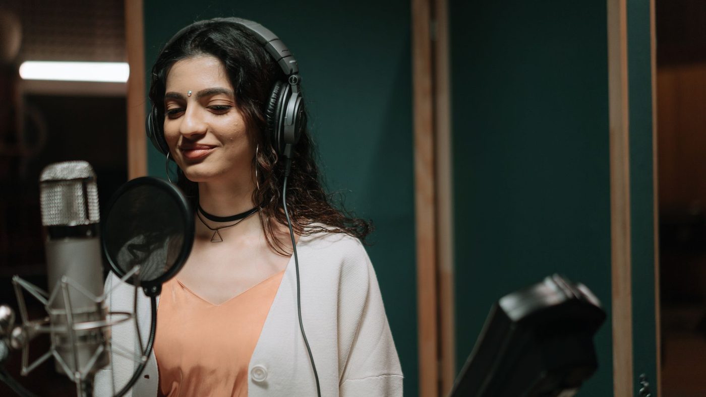 Woman singing into a microphone in a professional music studio wearing headphones.