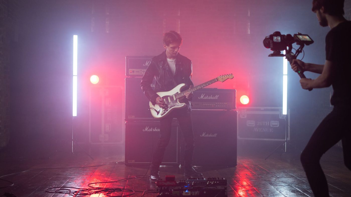Guitarist in a leather jacket performs in a studio setting with vibrant lighting, captured by a cameraman.