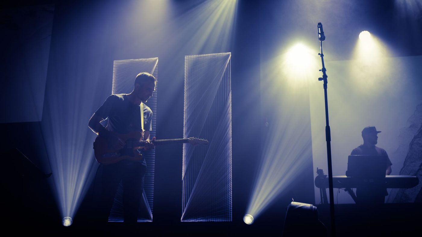 Anonymous male musician performing melody with guitar on stage near microphone and DJ in dark club with spotlights