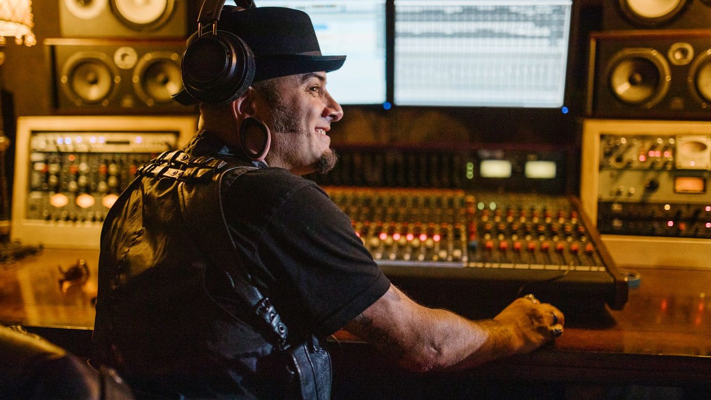 A person working in a recording studio wearing headphones and a hat, using audio equipment.